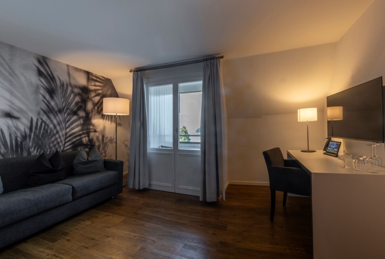Una habitación de hotel moderna con un sofá gris, una mesa de comedor con sillas, un espejo grande y una ventana con cortinas.