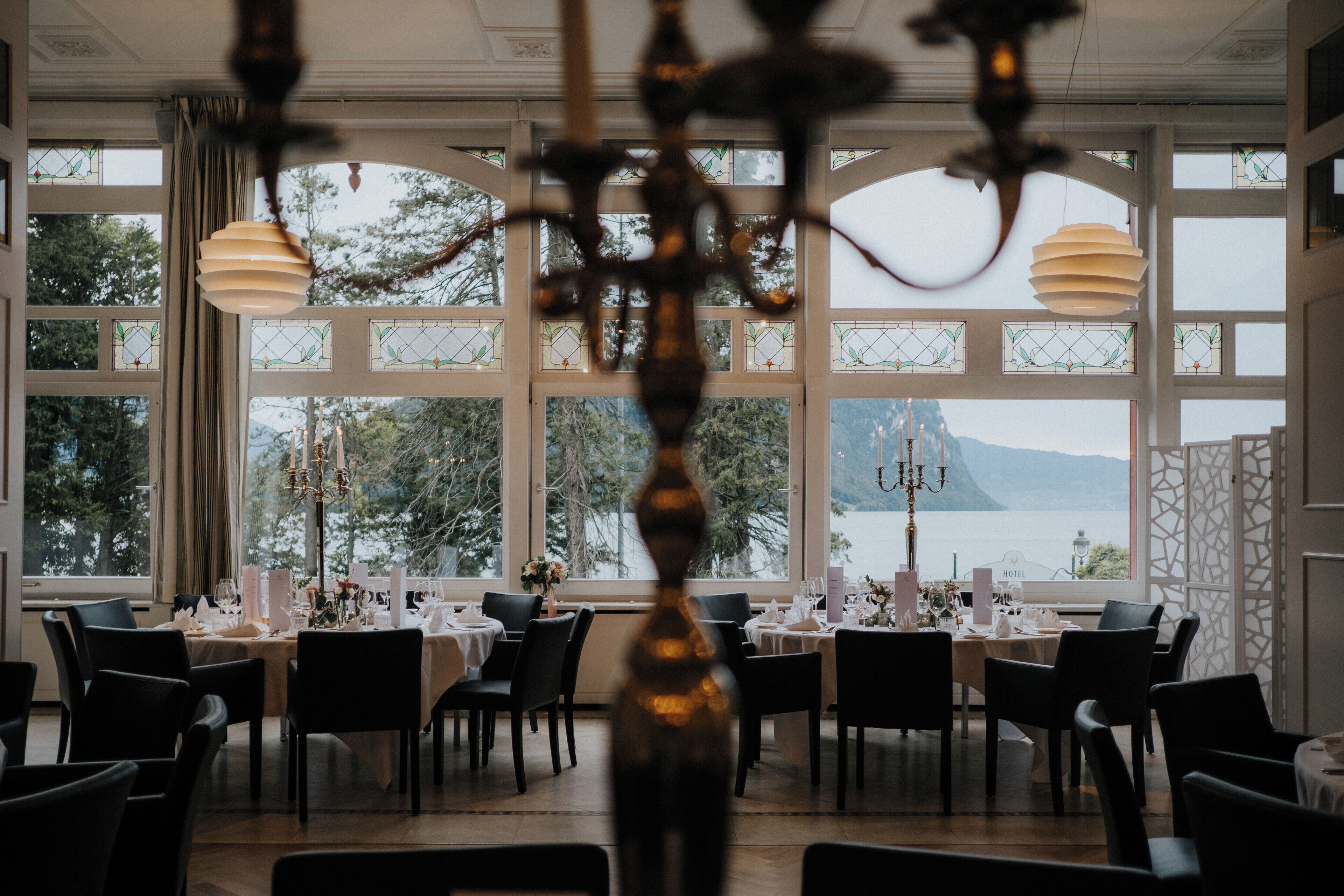 Un comedor con mesas dispuestas para una comida, ventanas amplias con vistas al agua y montañas, y una araña en primer plano.