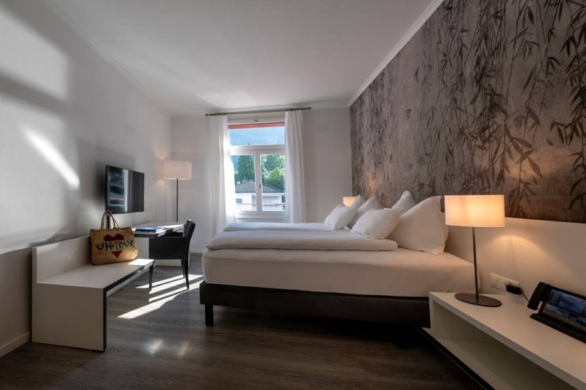 Habitación de hotel moderna con cama, mesitas de noche, escritorio y una silla. Se ve vegetación a través de la ventana.