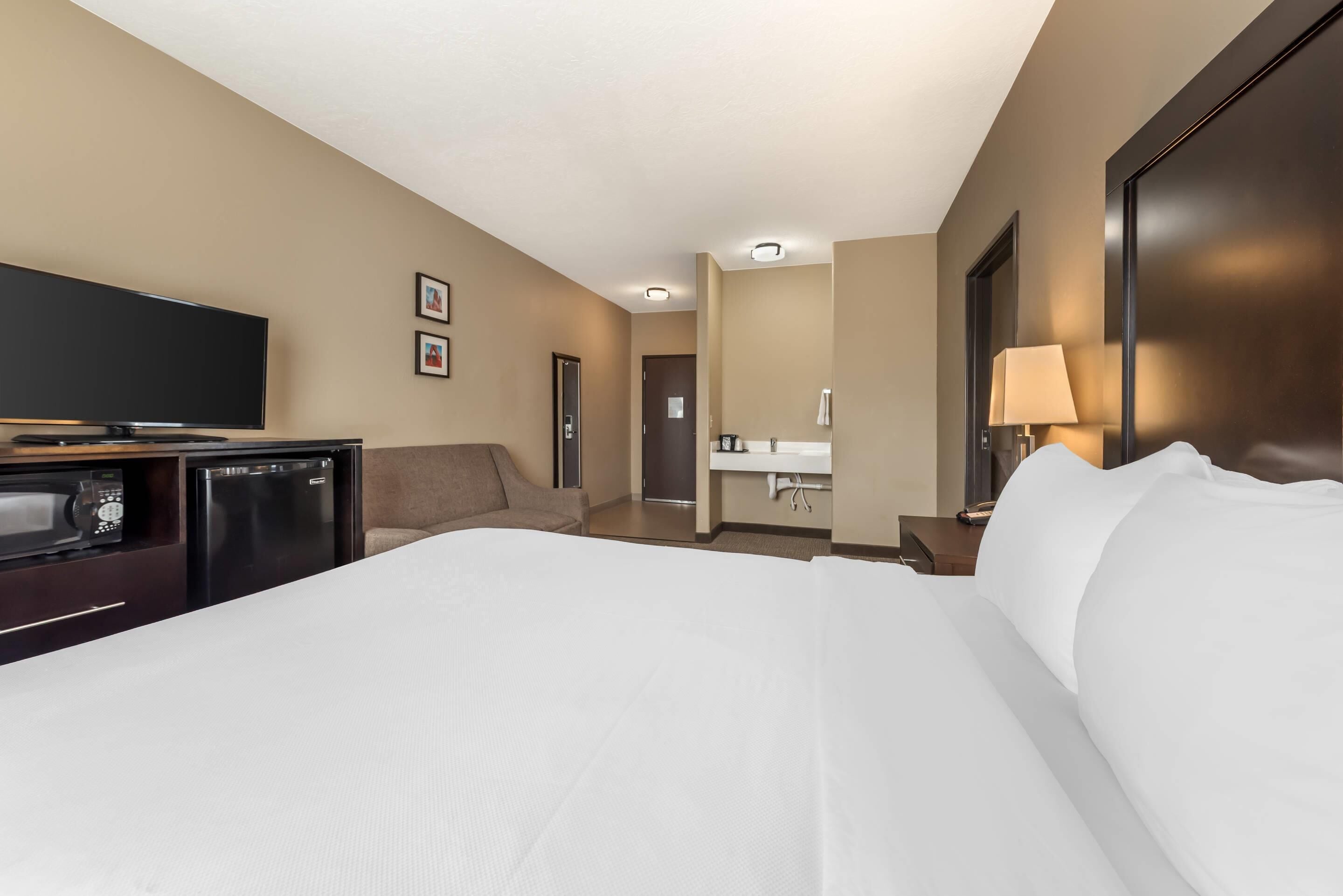 Habitación de hotel con una cama grande, televisor de pantalla plana, mini refrigerador, escritorio y baño visible a través de una puerta abierta.