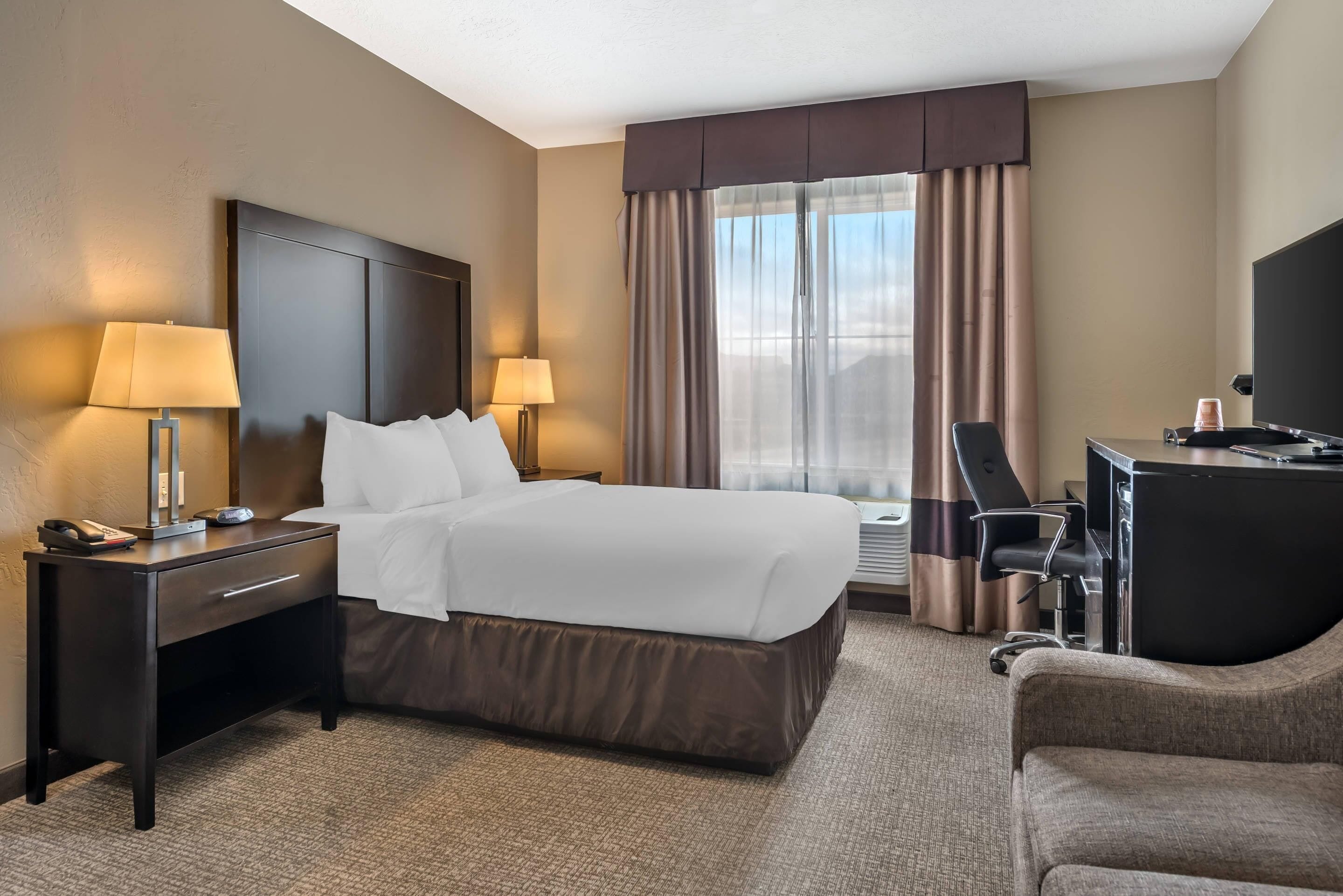 Quarto de hotel com uma cama grande, duas mesas de cabeceira com luminárias, uma escrivaninha com cadeira e uma janela ampla com cortinas.