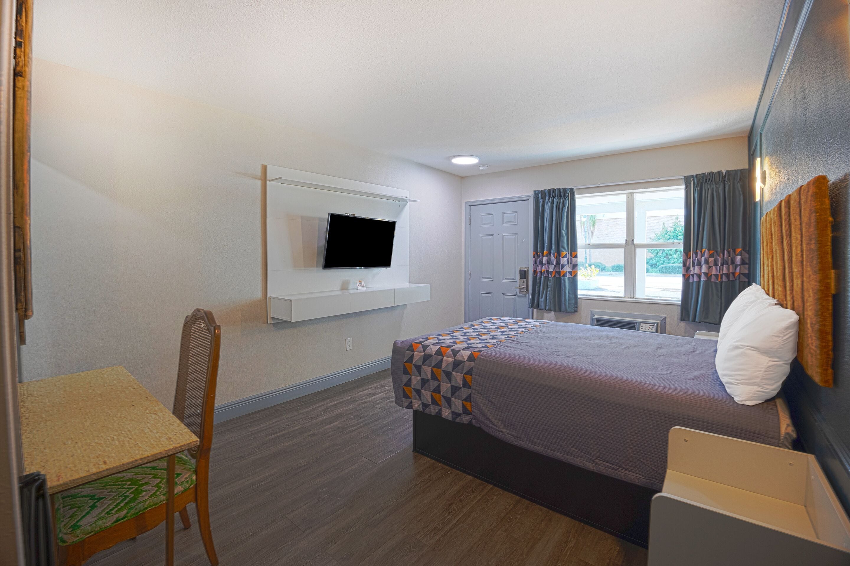 Una habitación de hotel con cama, televisor montado en la pared, una silla y una ventana con cortinas.