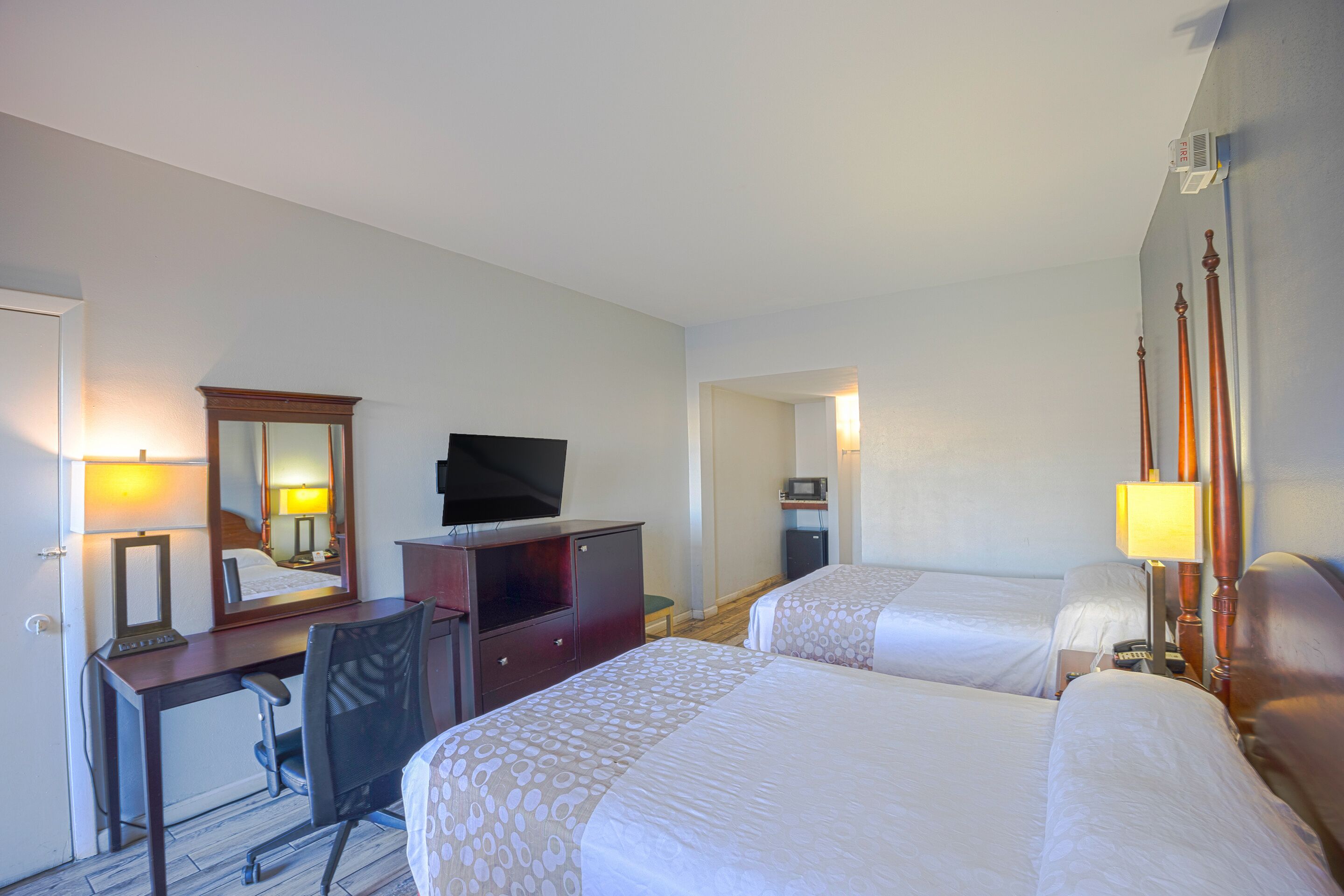 A hotel room with two beds, a desk with a chair, a television, a mirror, and a bedside table with a lamp.