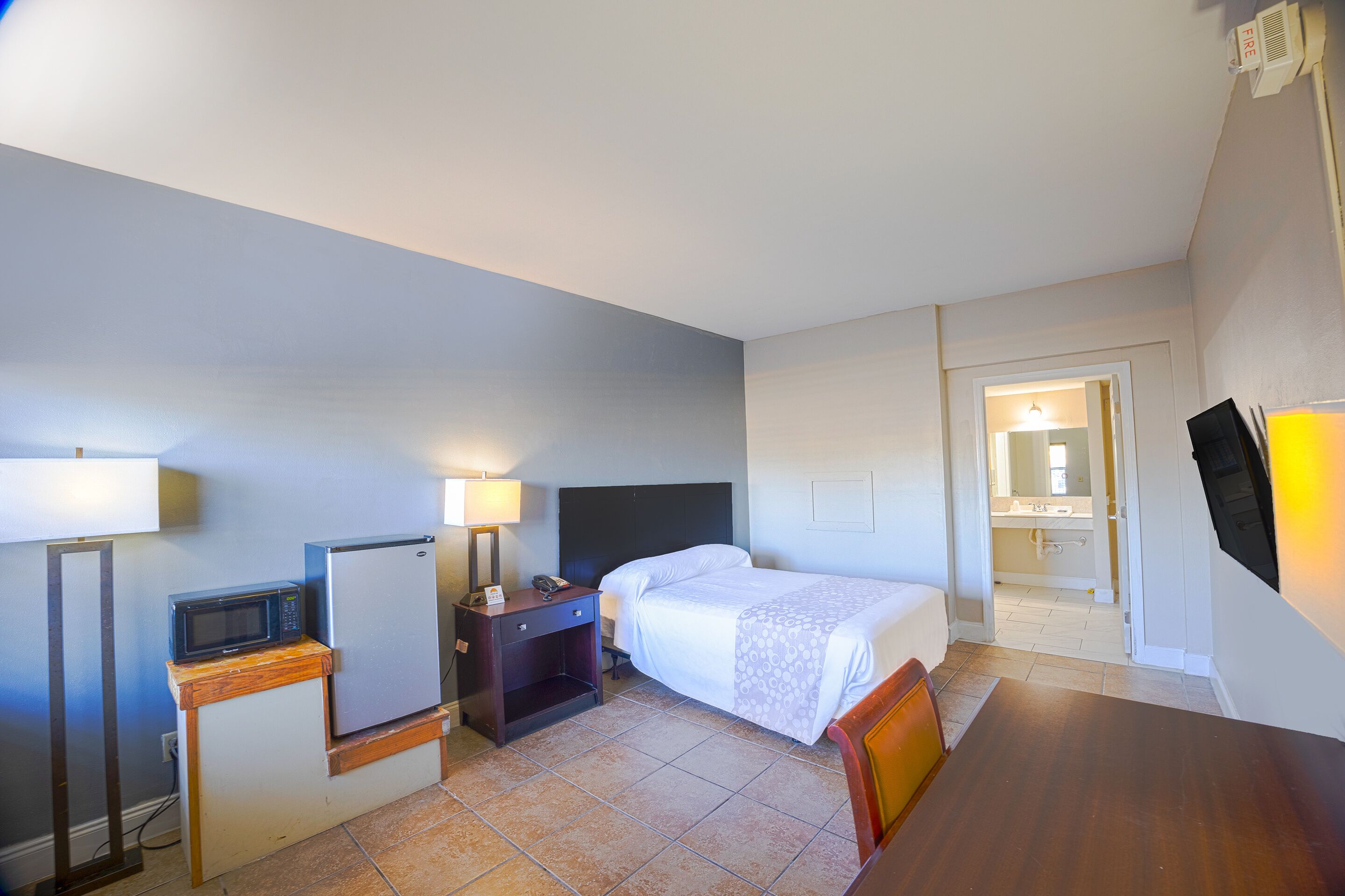 A hotel room with a bed, a desk, a chair, a microwave, a refrigerator, a nightstand, and a bathroom visible through an open door.