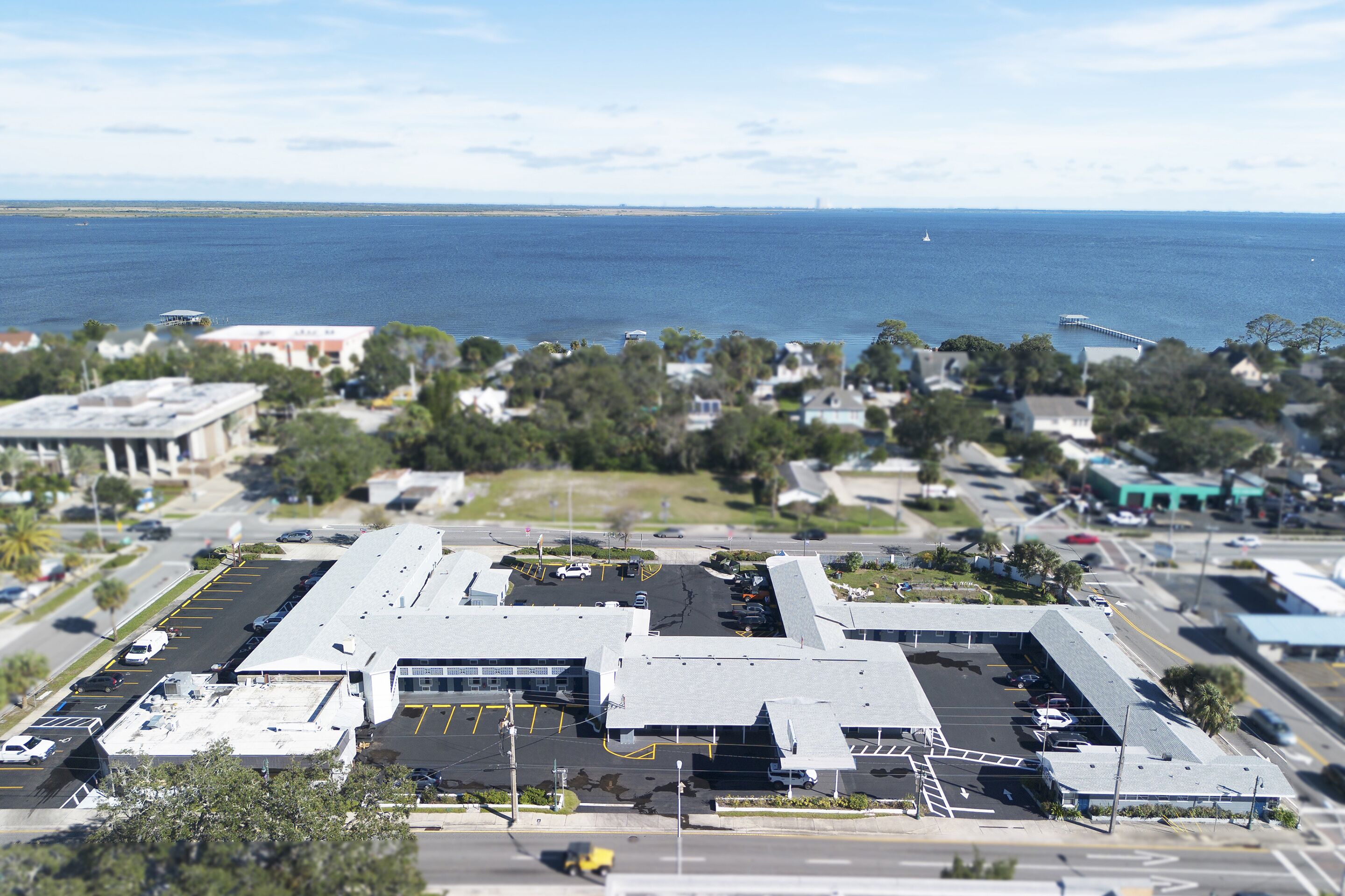 Vista aérea de um edifício comercial com estacionamento, rodeado por árvores e vista para o oceano.