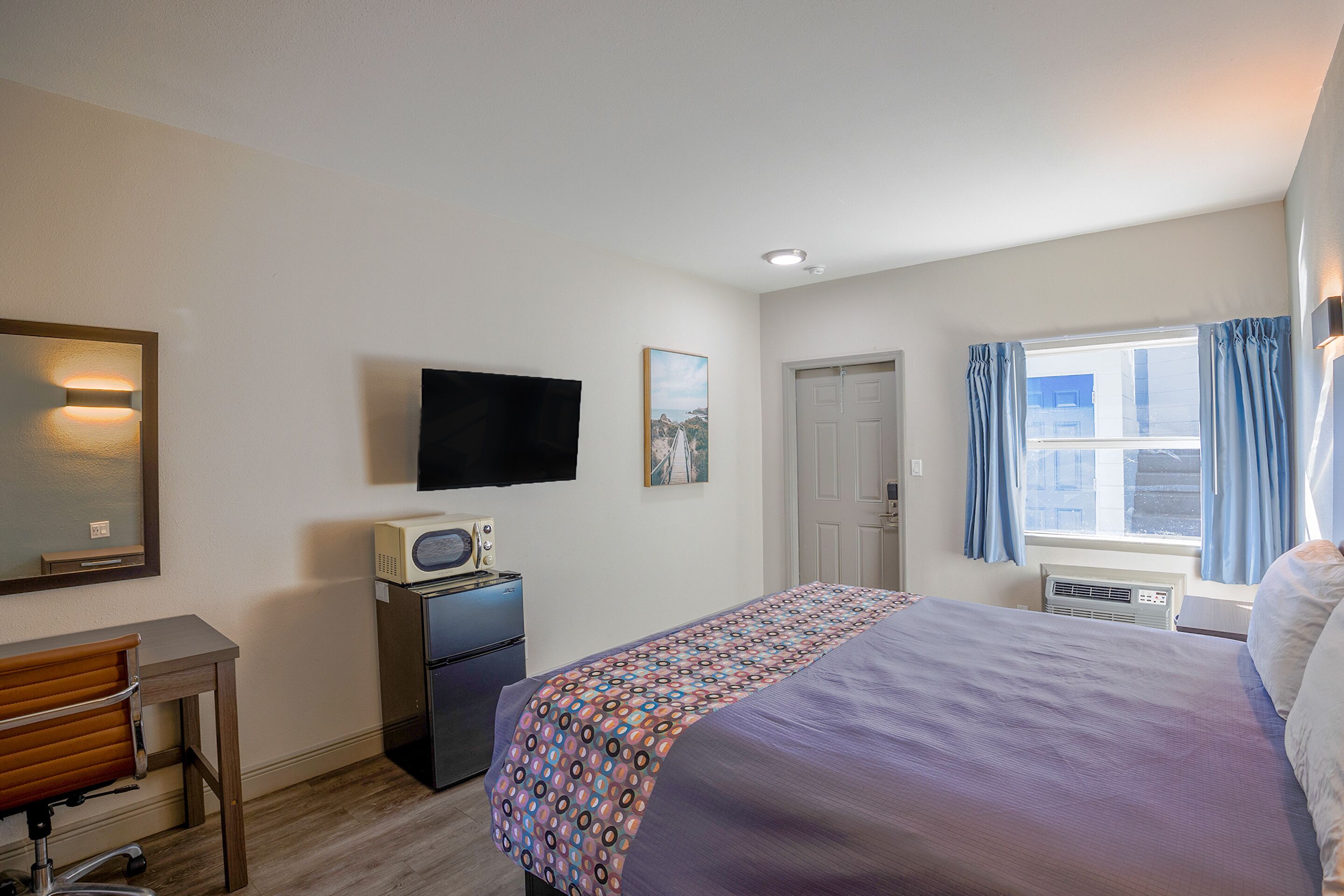 Ein Hotelzimmer mit Bett, Schreibtisch mit Stuhl, Fernseher, Kühlschrank, Fenster mit Vorhängen und einer Tür.