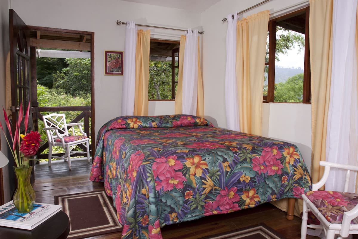 A bedroom with a bed, a chair, a window with curtains, and a view of greenery outside.