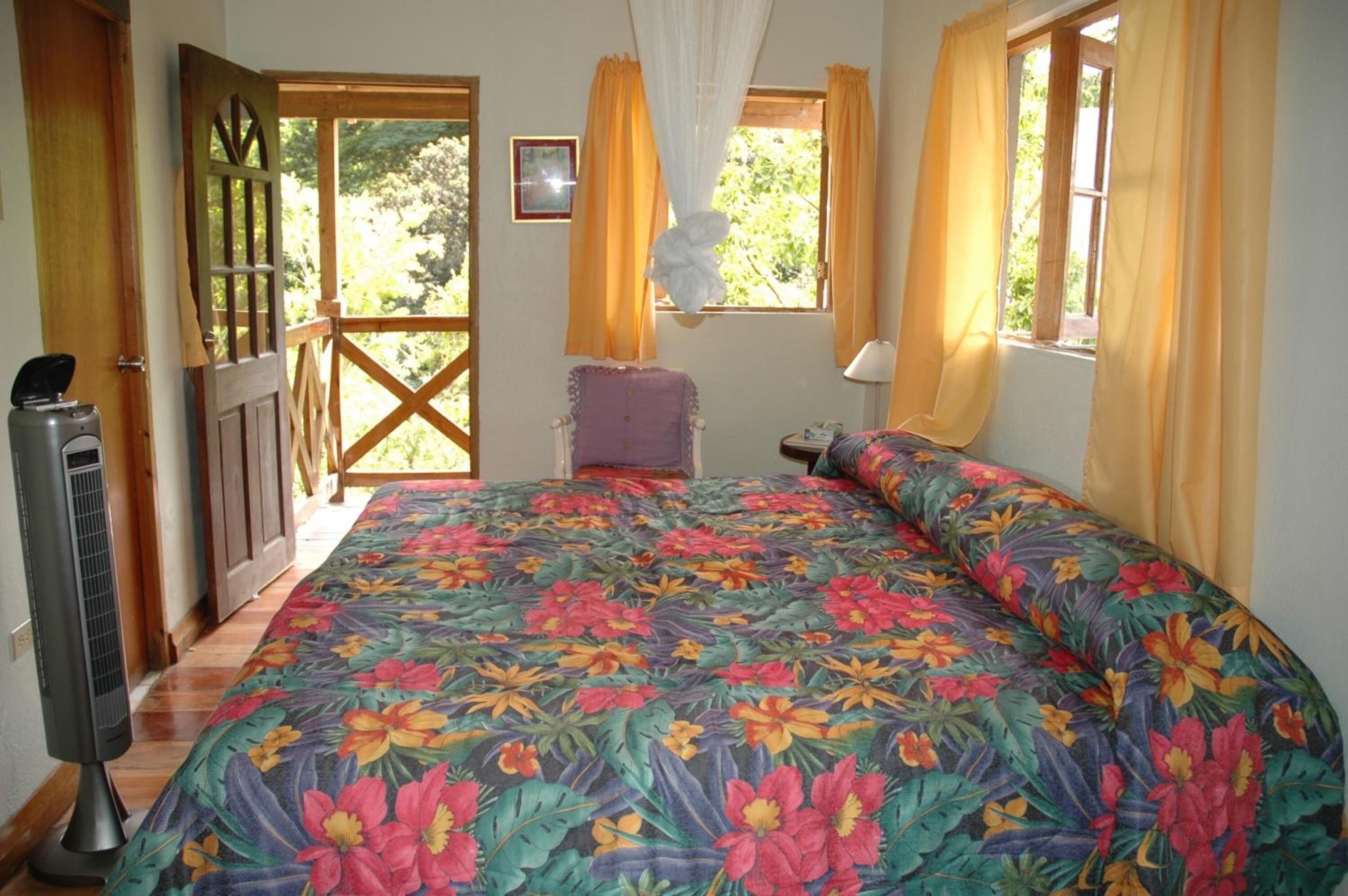 Un dormitorio con colcha estampada, puerta de madera, ventana con cortinas y una mesita de noche.