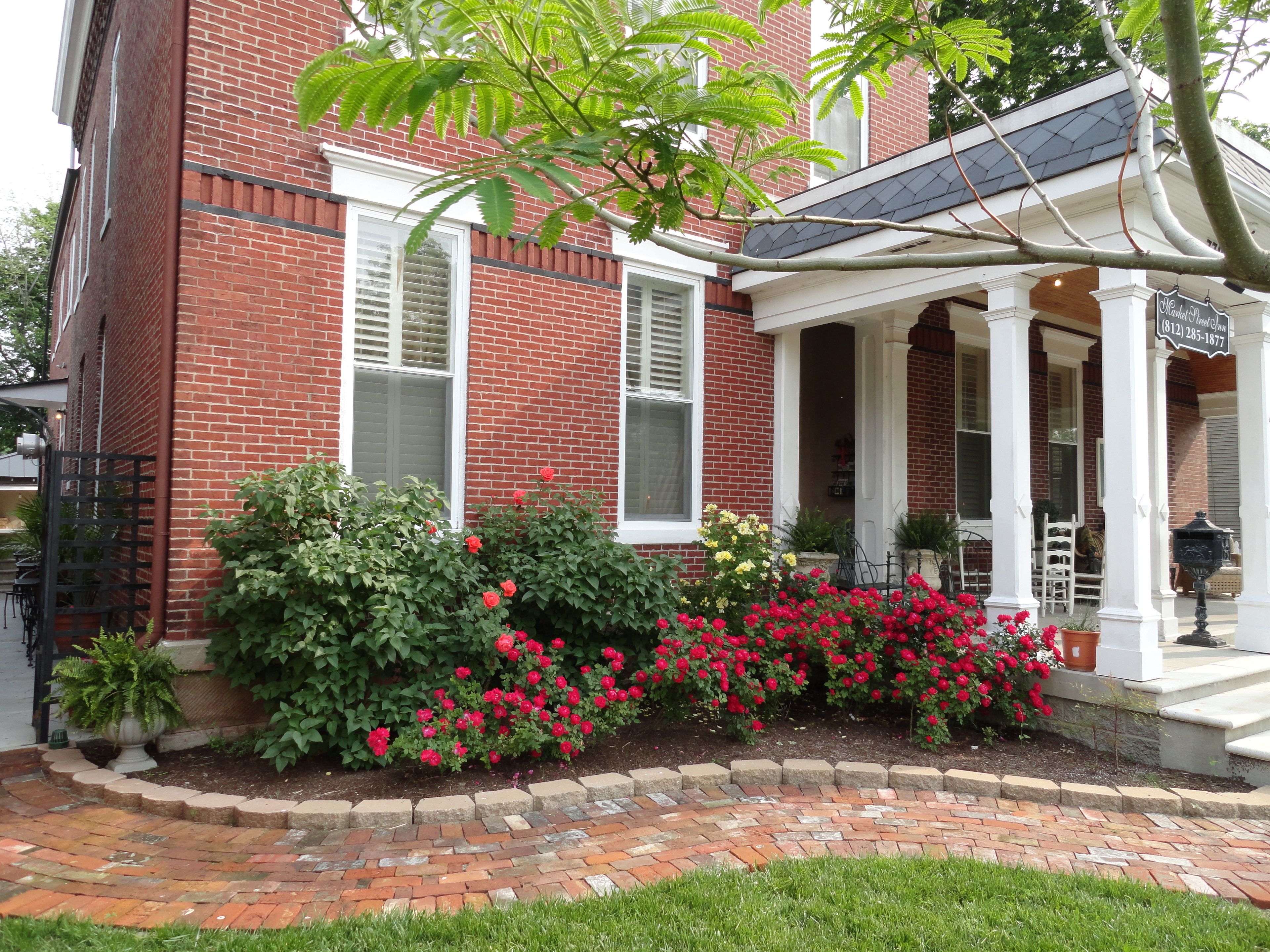 A red brick house with a covered porch, a brick pathway, and red flowers in the garden.