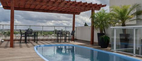 A rooftop pool with wooden deck, a pergola, and a glass enclosure with plants.
