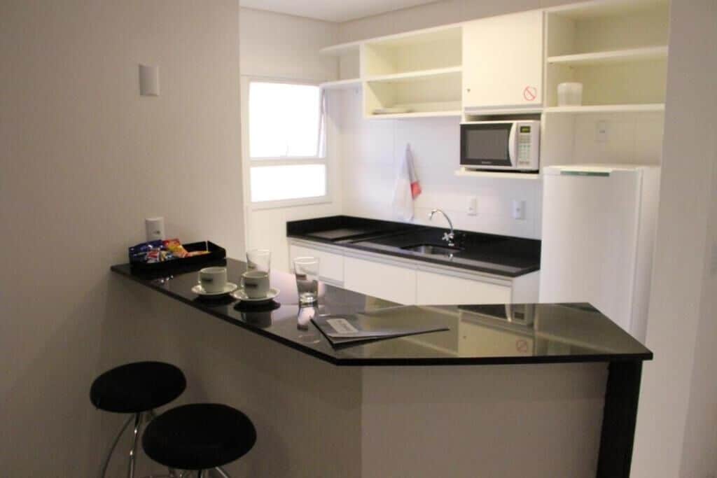 Una cocina moderna con encimera negra, armarios blancos, microondas y refrigerador. Hay dos taburetes negros y una pequeña mesa con tazas y un vaso.