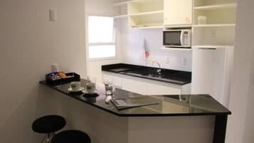 Una cocina moderna con encimera negra, armarios blancos, microondas y refrigerador. Hay dos taburetes negros y una pequeña mesa con tazas y un vaso.