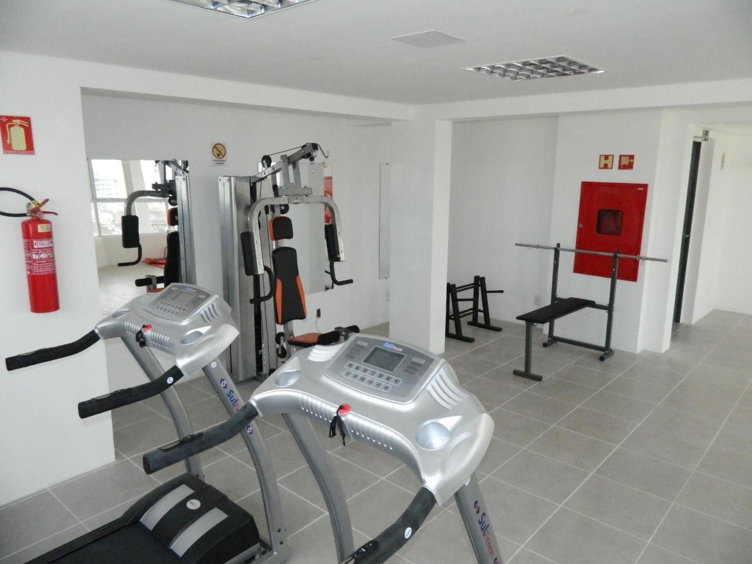 Un gimnasio con cintas de correr, máquinas de pesas y un extintor rojo en la pared.