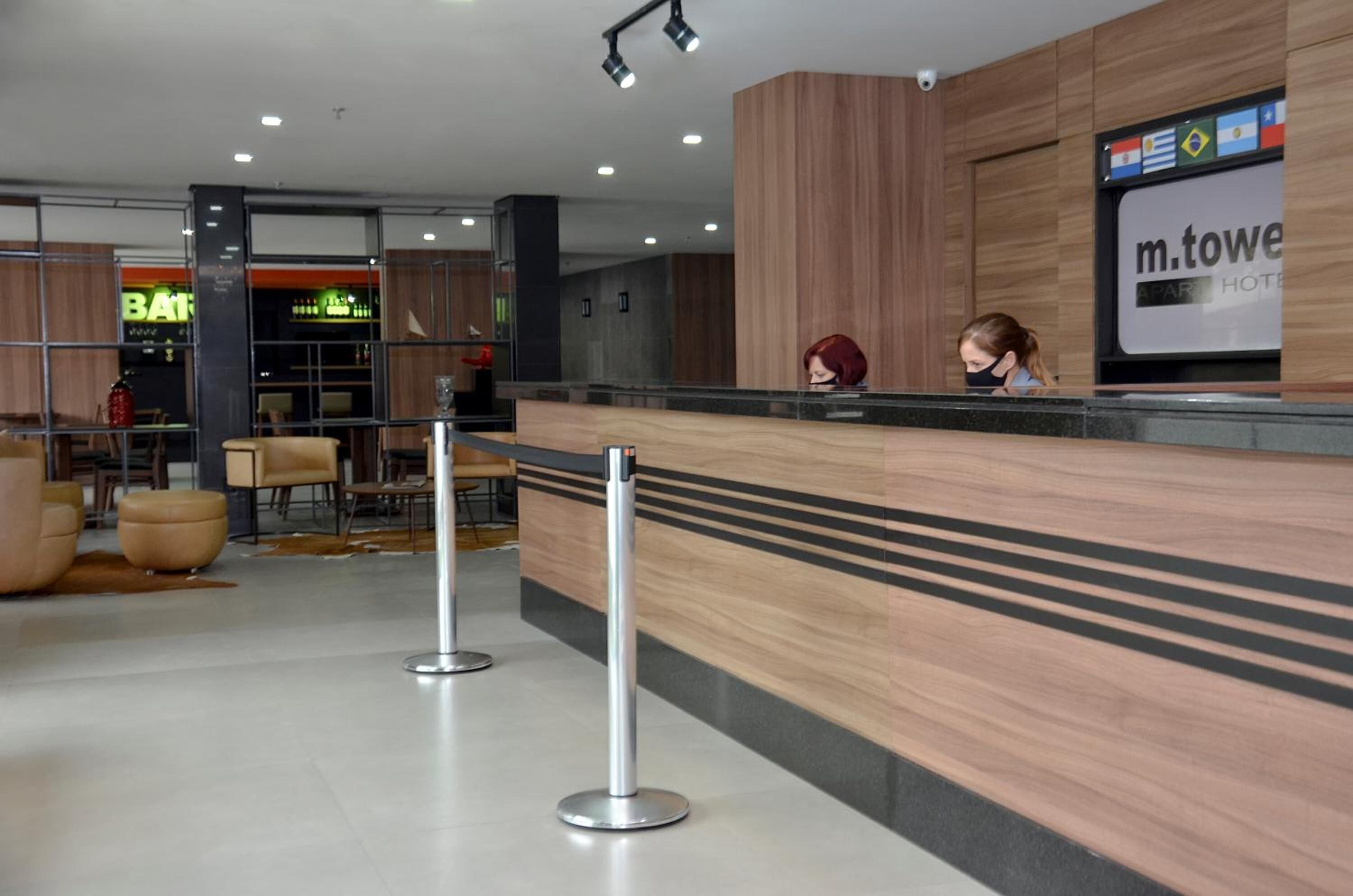Un área de recepción moderna de hotel con un mostrador de madera, una pantalla digital y una zona de estar con sillones y otomanas de cuero.