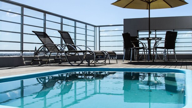Un área de piscina con sillones de descanso y una sombrilla, rodeada por una terraza de madera y una barandilla de cristal.