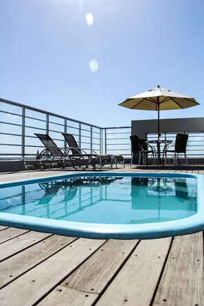 A pool area with lounge chairs and an umbrella, surrounded by a wooden deck and a glass railing. - M. Tower Hotel (Pelotas)