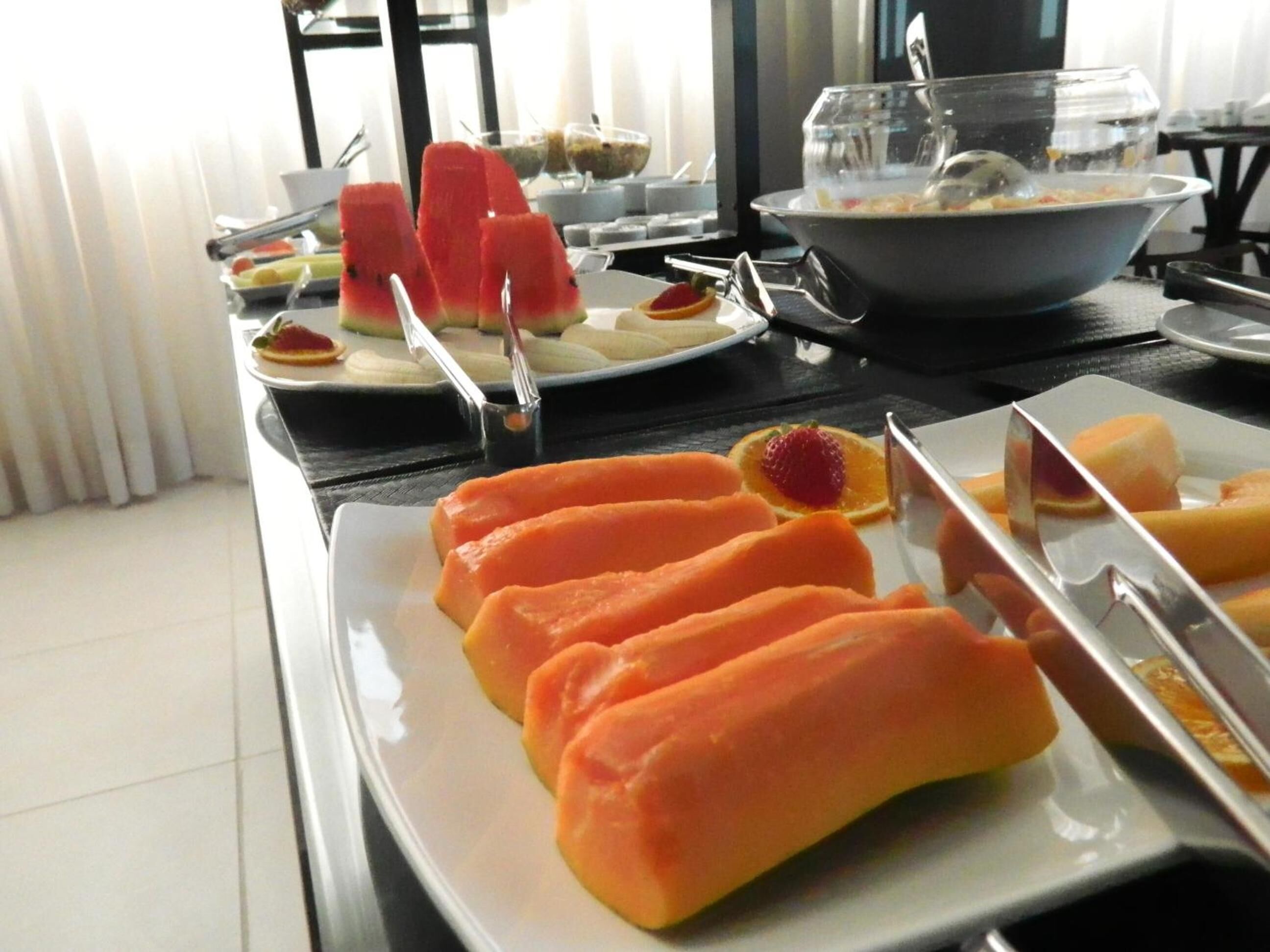 Uma mesa de buffet com diversos alimentos, incluindo melancia e rodelas de laranja, e uma tigela de sopa.