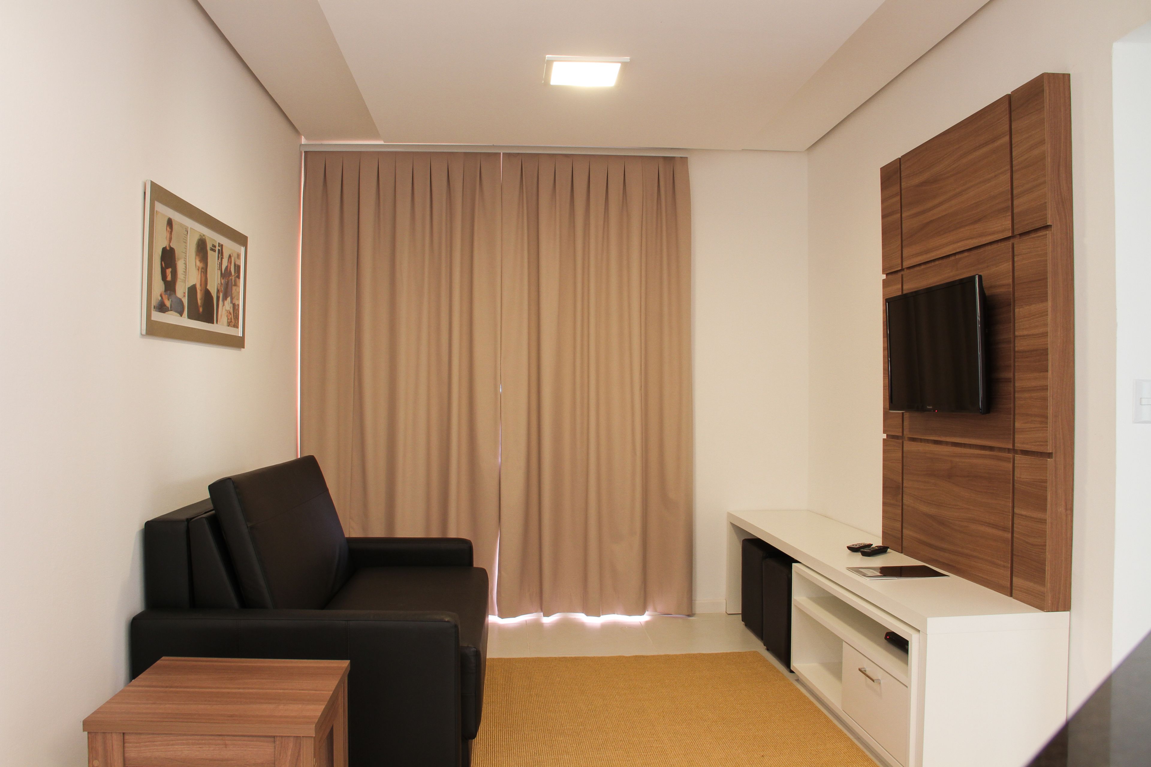 A modern living room with a black sofa, a wooden coffee table, a TV mounted on a wooden cabinet, and a large window with curtains.