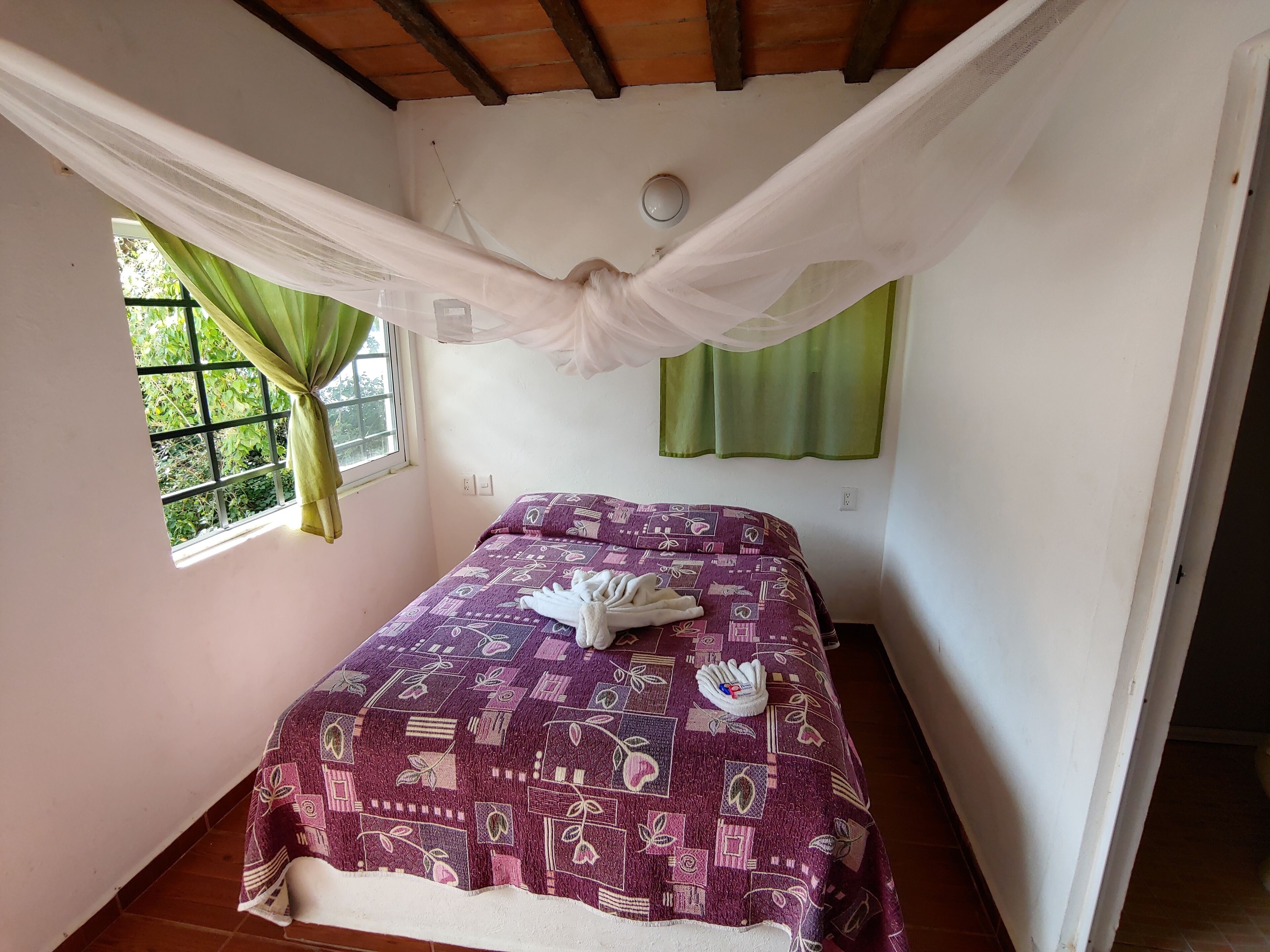 A bedroom with a bed covered in a patterned bedspread, a window with green curtains, and a ceiling with wooden beams.