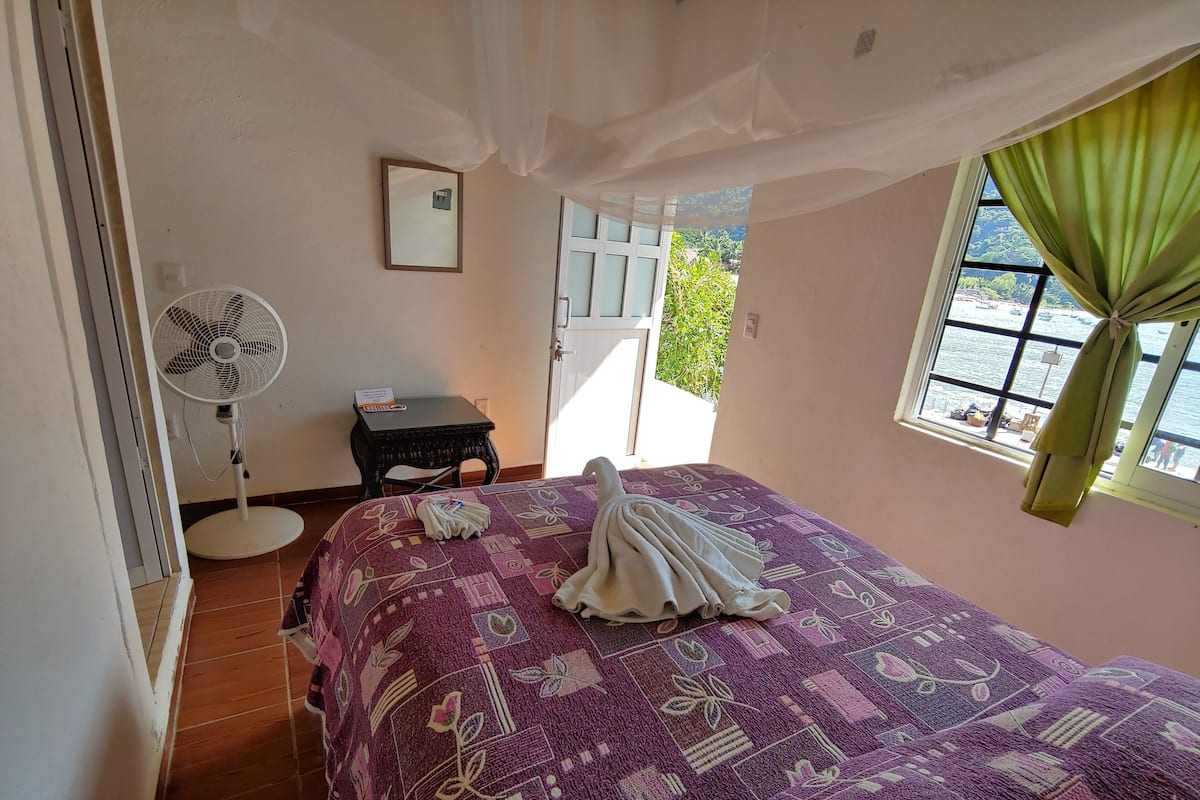 Ein Schlafzimmer mit einem Bett, einem Standventilator, einem kleinen Tisch und einem Fenster mit Vorhängen.