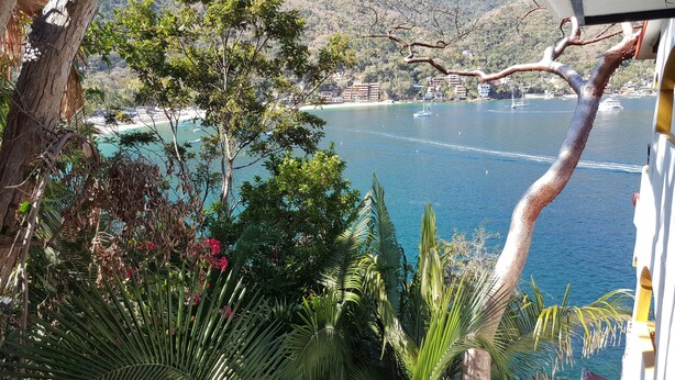 A coastal view with a clear blue sea, a sandy beach, and a backdrop of buildings and hills.
