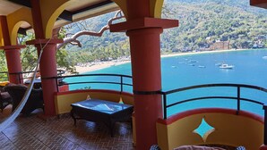 A balcony with a view of a beach and boats, featuring red pillars and wicker furniture.