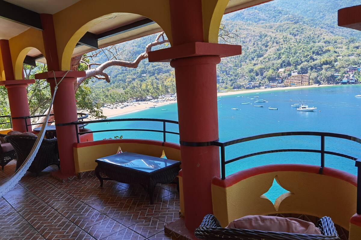 Ein Balkon mit Blick auf einen Strand und Boote, mit roten Säulen und Korbmöbeln.