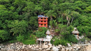 A red building with multiple balconies, surrounded by lush greenery and a rocky shoreline.