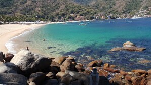 A coastal beach with clear turquoise waters, large rocks in the foreground, and a hilly backdrop with buildings.
