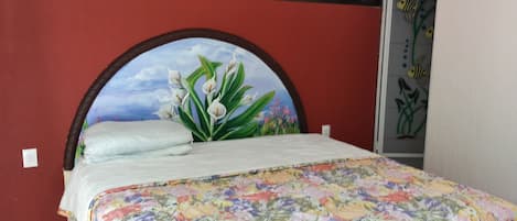 A bed with a floral bedspread, a headboard with a painted landscape, and a red wall.