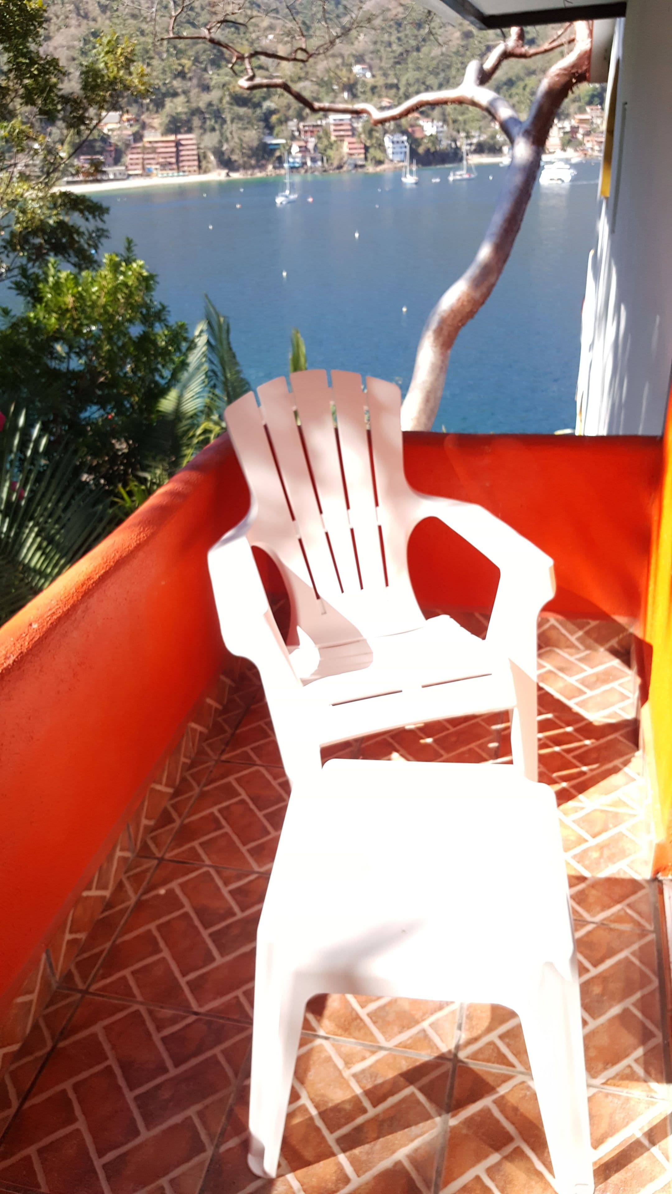 A white plastic chair on a tiled balcony overlooking a body of water with boats and buildings in the distance.