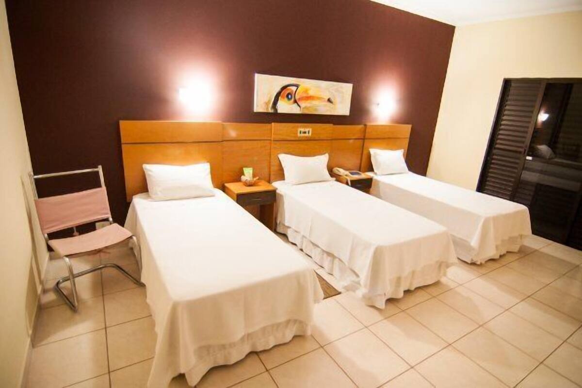 A hotel room with three single beds, a wooden headboard, a painting of a bird, and a window with blinds.