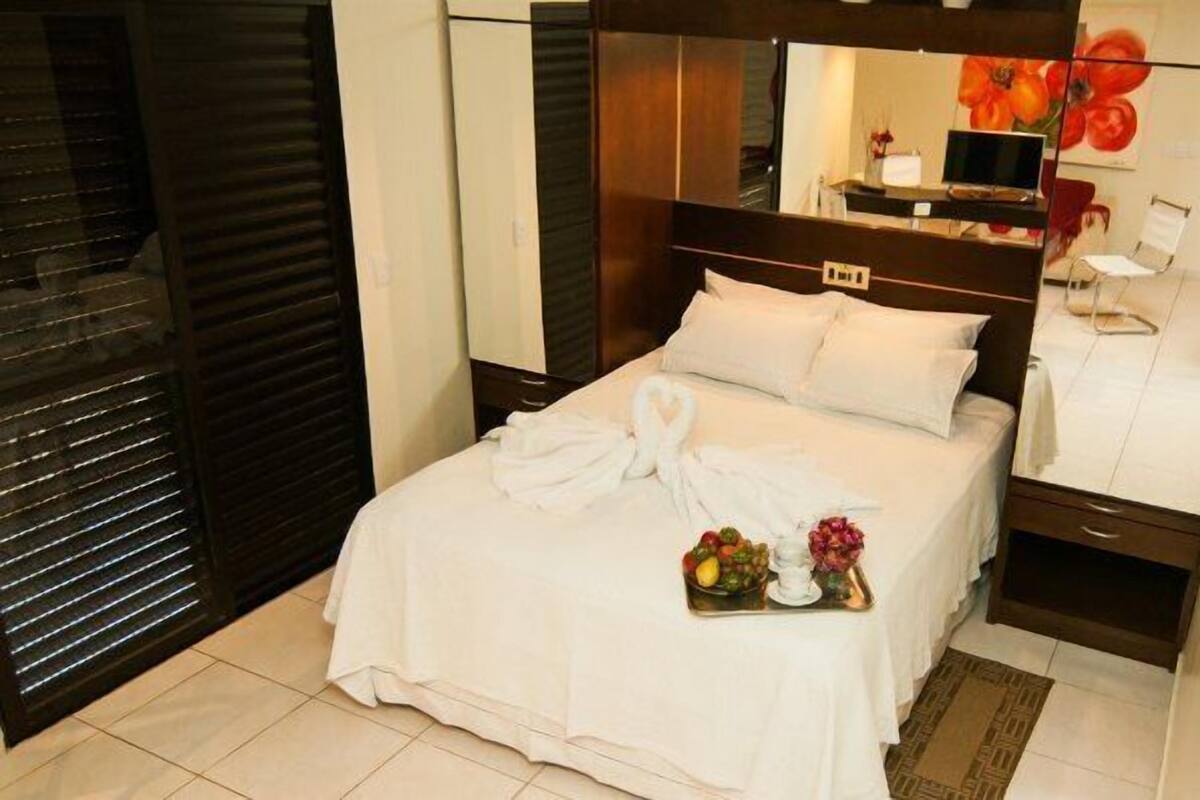 A neatly made bed with a towel and fruit basket, a bedside table with a lamp, and a mirror above the bed.