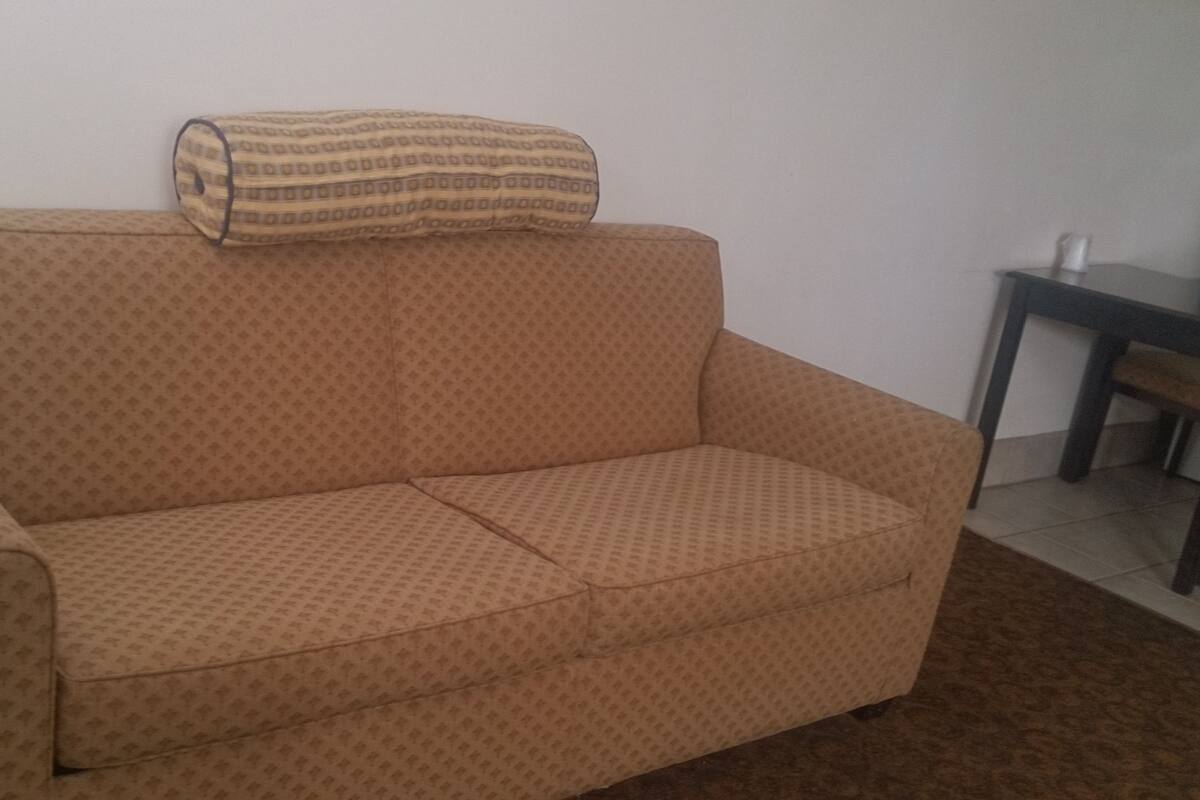 A beige sofa with a patterned fabric, a rolled-up blanket on top, and a small table with a white object on it.
