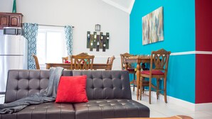Een woonkamer met een donker leren bank, een rode kussen, een houten eettafel met stoelen, en een keuken met een koelkast en magnetron.