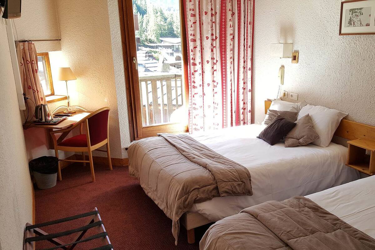 A hotel room with two beds, a desk, a chair, a lamp, and a window with curtains.