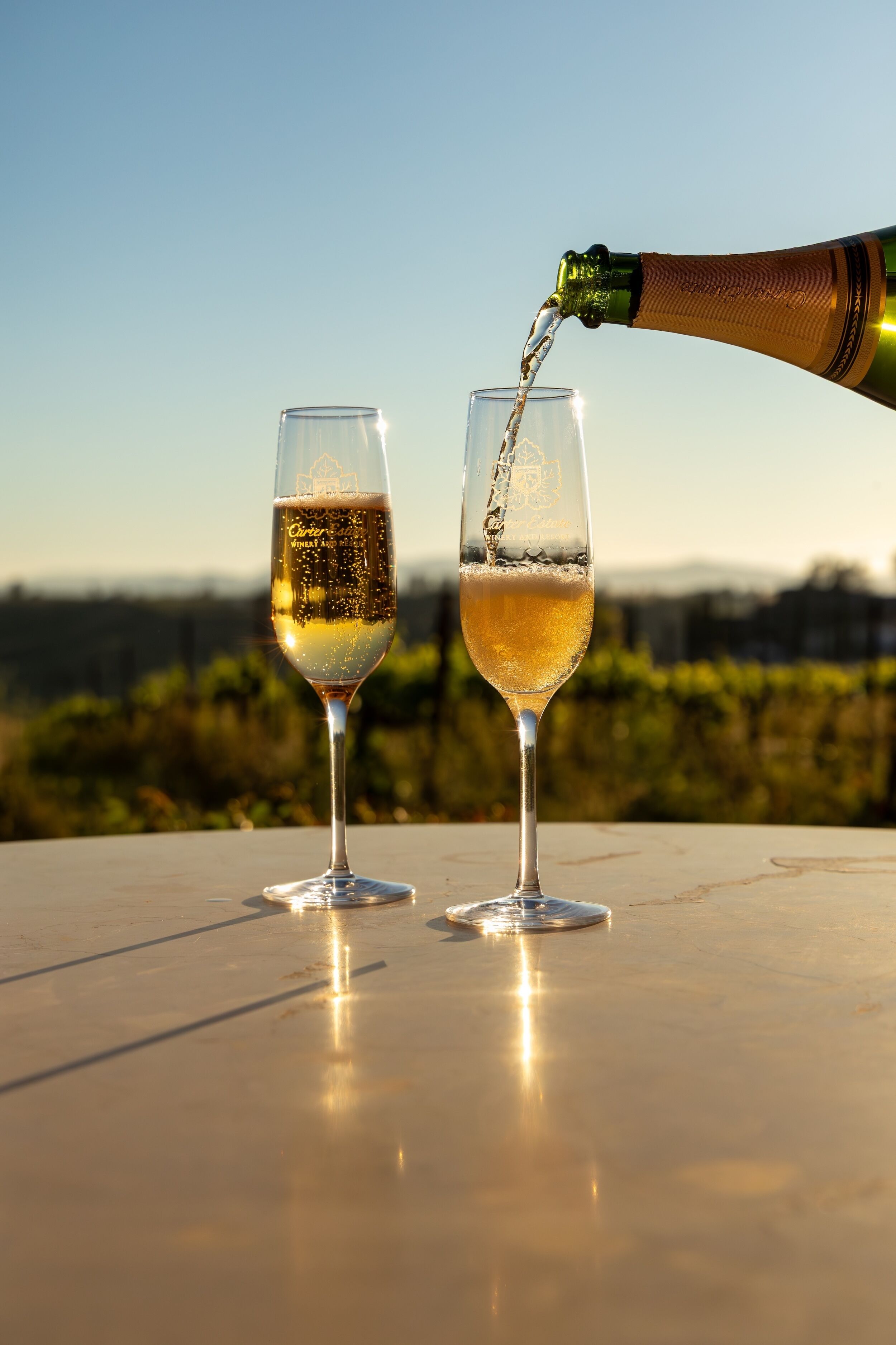 Two champagne glasses on a table with a bottle pouring into one.