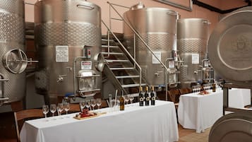 A wine tasting room with stainless steel fermentation tanks, a white table with wine glasses and bottles, and a staircase leading to an upper level.