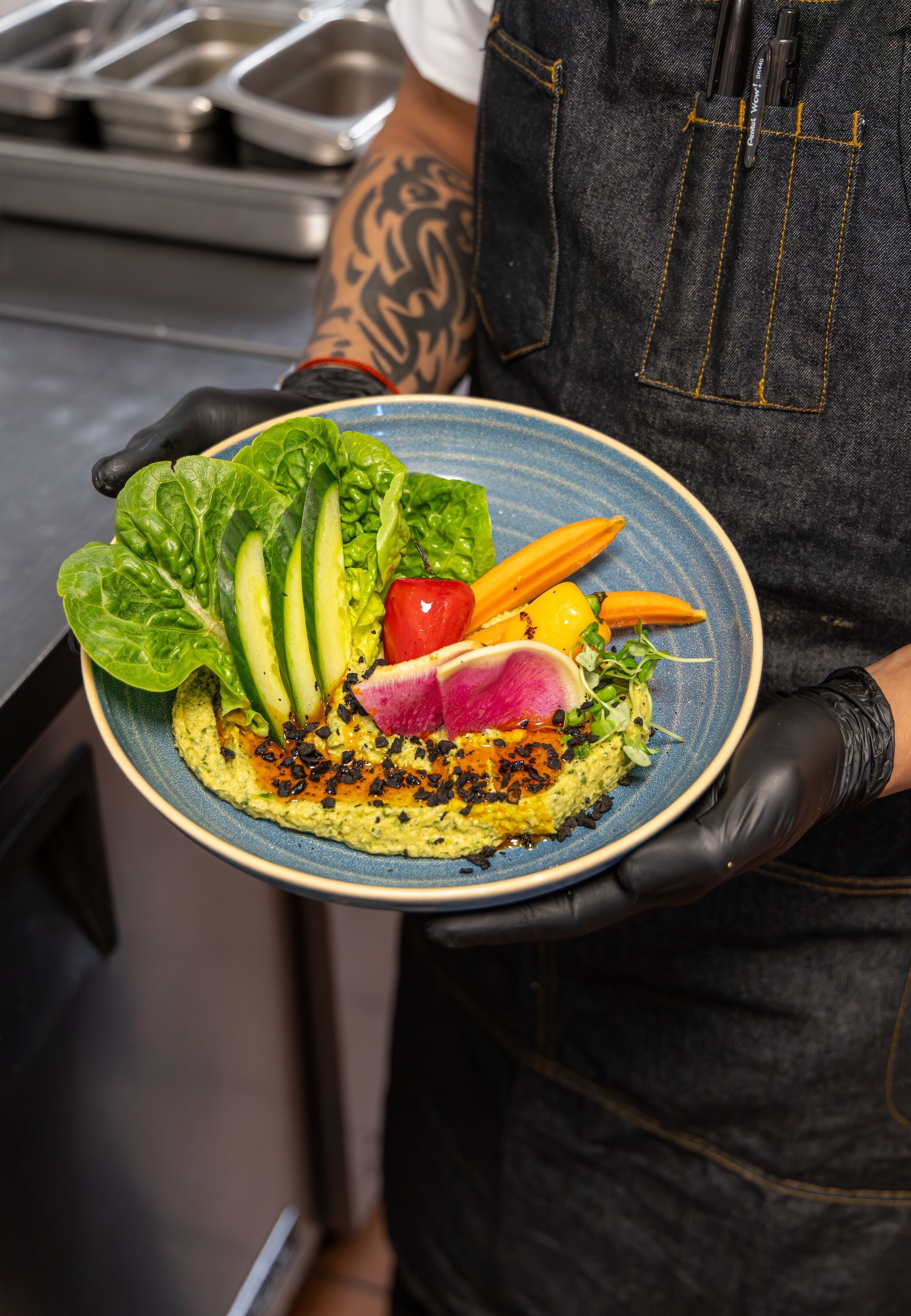 Une personne tenant une assiette de salade, portant un tablier noir et ayant un tatouage sur le bras.