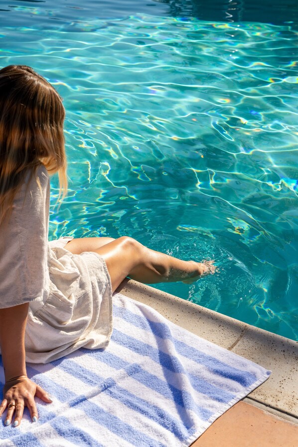 Uma pessoa sentada à beira de uma piscina com água cristalina, uma toalha e borda de azulejos.