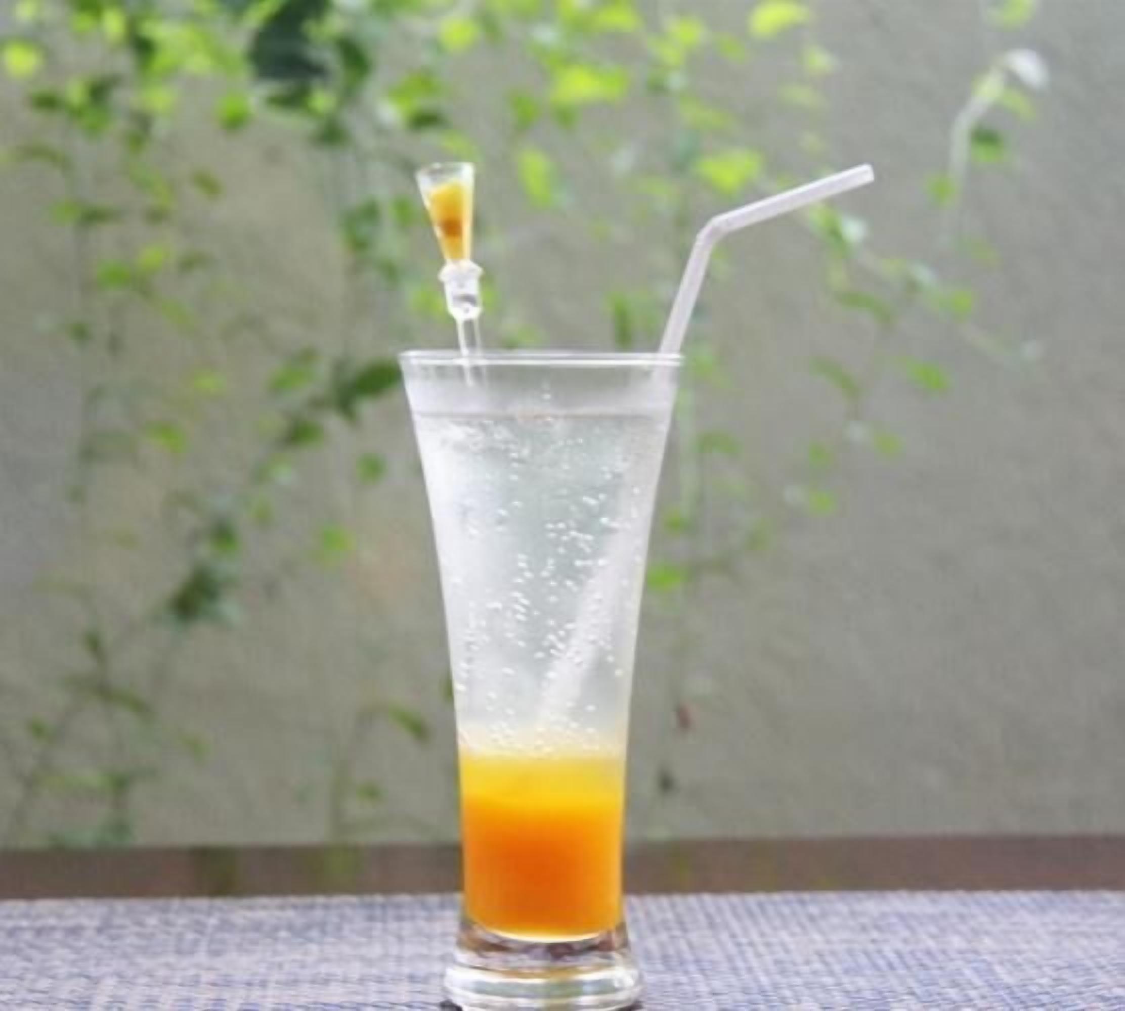 A tall glass with a straw and a slice of lemon on the rim, filled with a yellow-orange liquid, likely juice, against a backdrop of greenery.