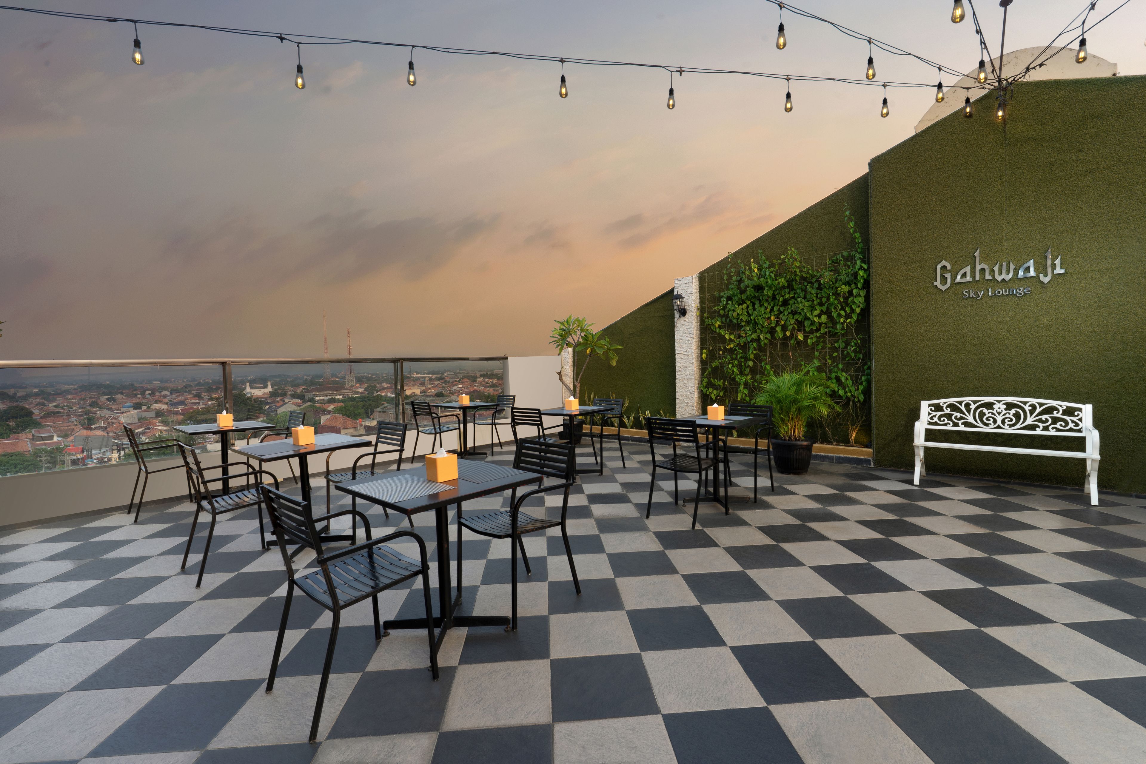 Un espace extérieur aménagé avec des tables et des chaises, offrant une vue sur la ville, et un mur portant l’inscription « Gahwa J1 Sky Lounge ».