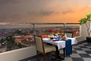 A dining table set with a vase of red flowers, candles, and a menu sign on a balcony overlooking a cityscape. - Hotel Namira Syariah Pekalongan (Pekalongan)