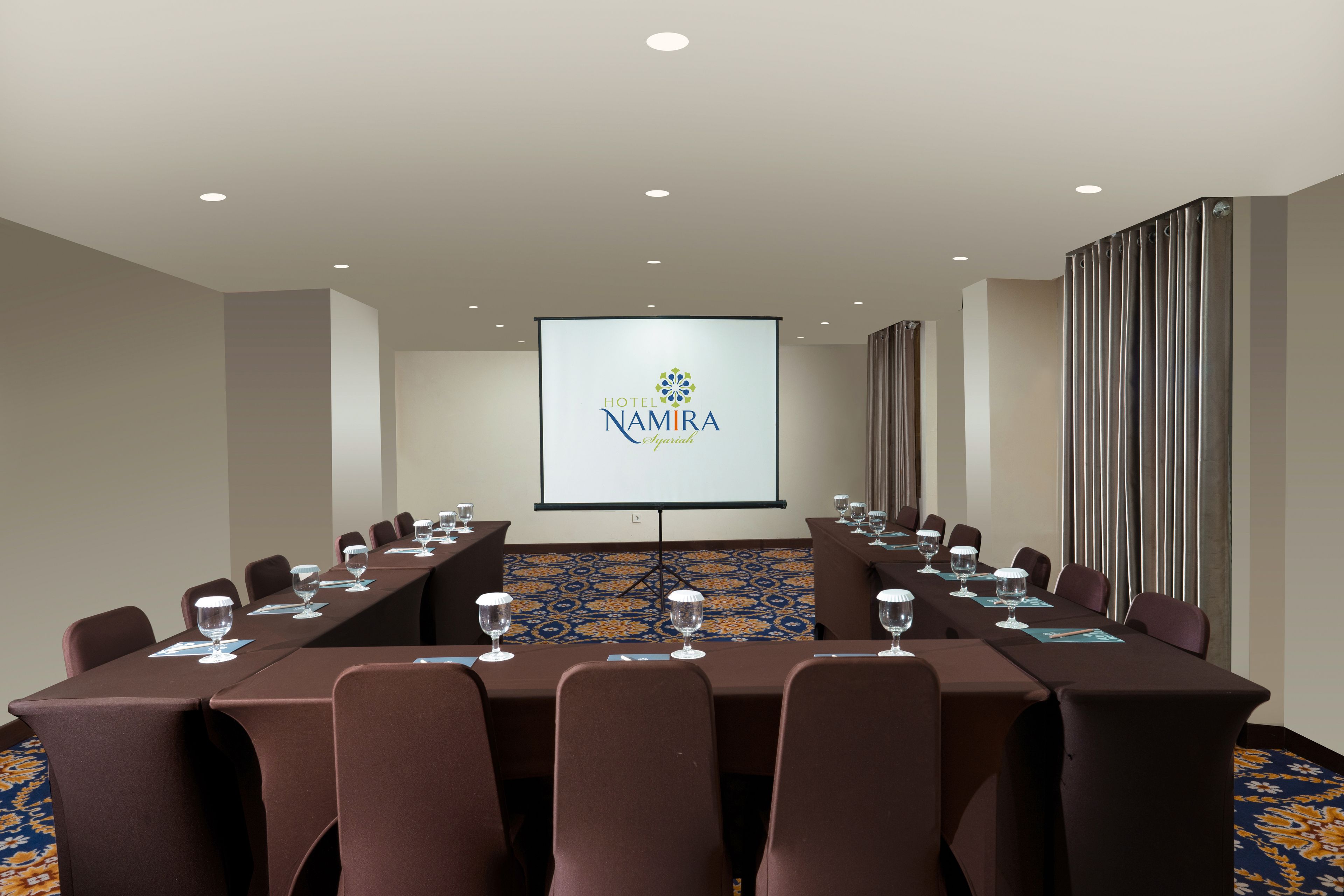 Un salón de conferencias con una larga mesa, sillas y una pantalla que muestra el nombre del hotel.