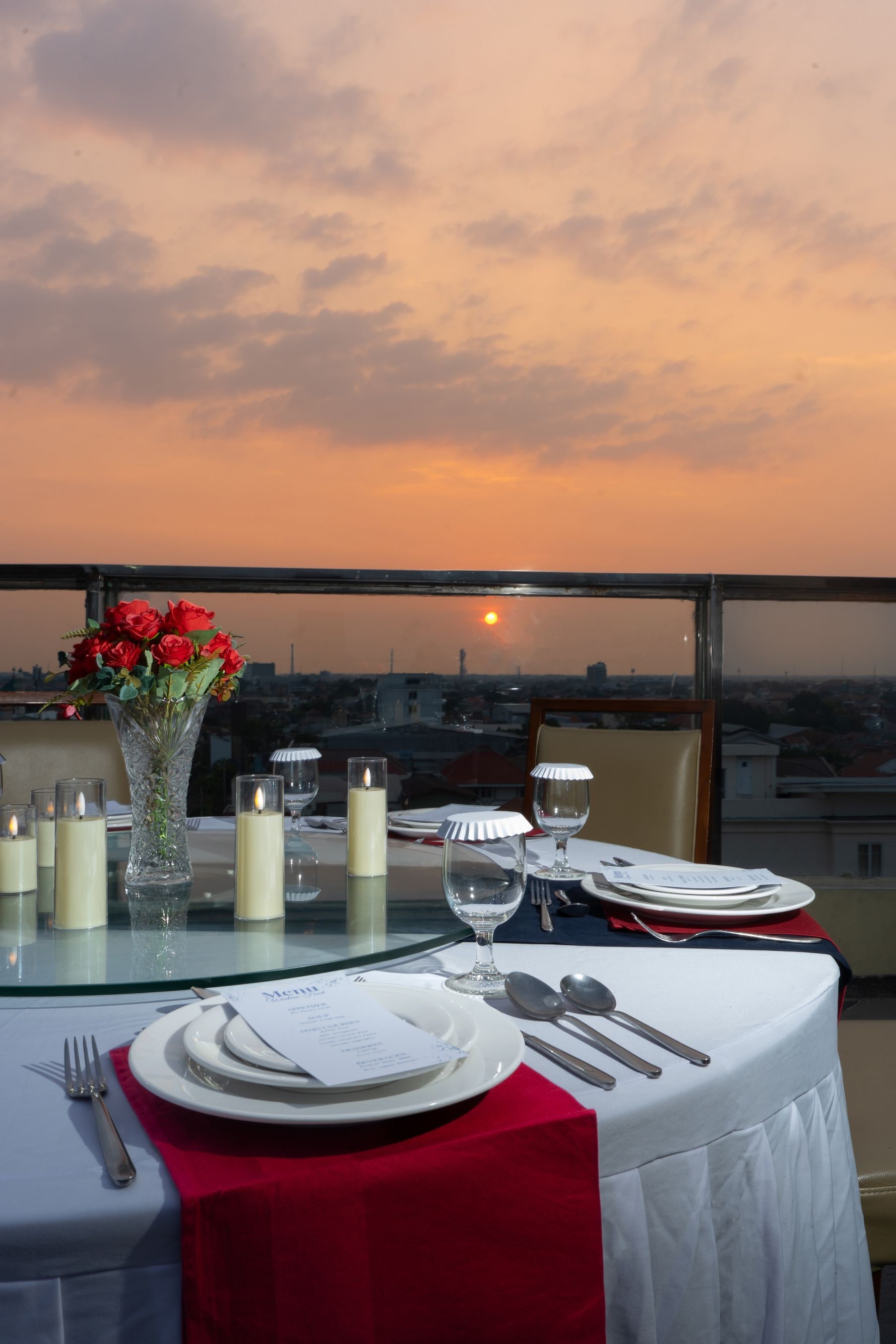 A table set for dining with candles, a vase of flowers, and a view of the sunset.