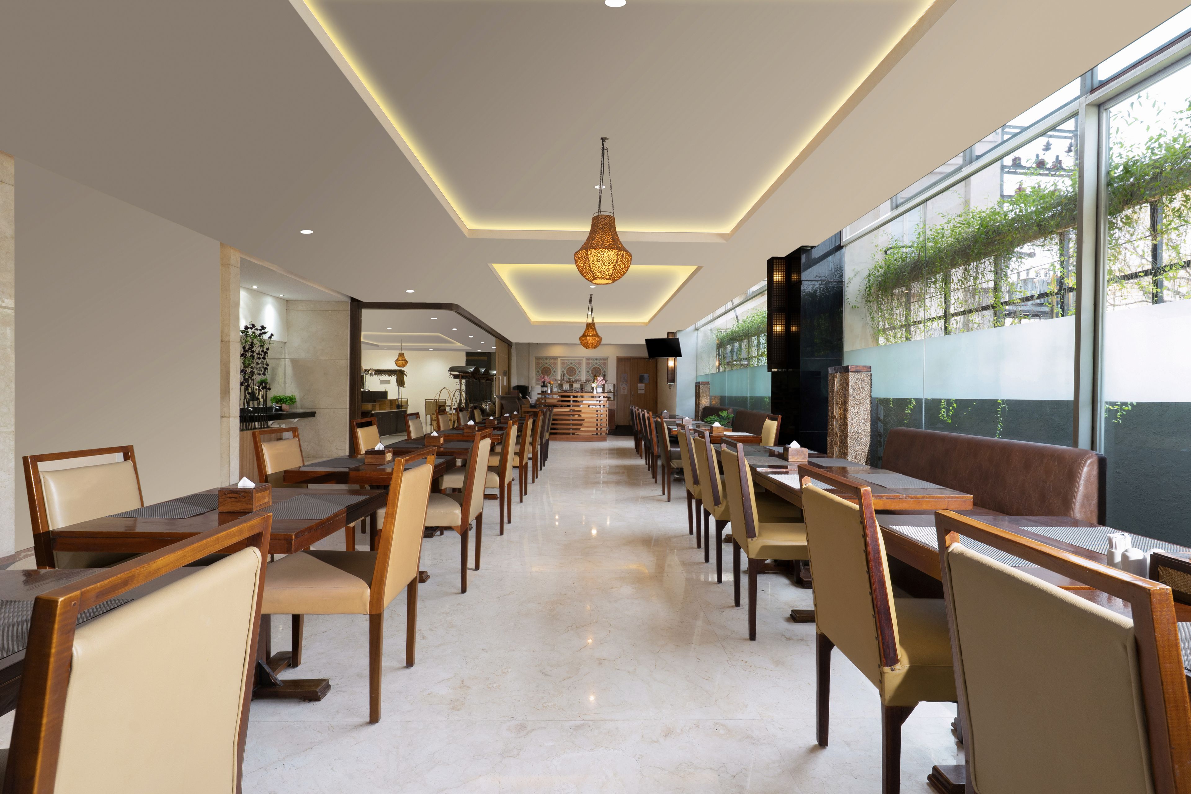 A spacious dining area with multiple tables and chairs, a modern chandelier, and large windows offering a view of greenery outside.