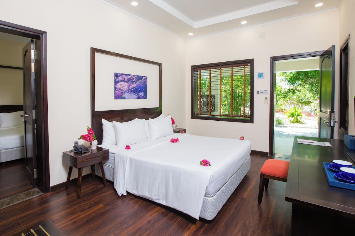 A hotel room with a large bed, a bedside table with a phone, a nightstand with a vase of flowers, a window with a view of greenery, and a desk with a chair and a tray with cups.