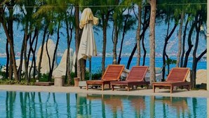 A poolside area with lounge chairs and umbrellas, surrounded by trees and overlooking a body of water.