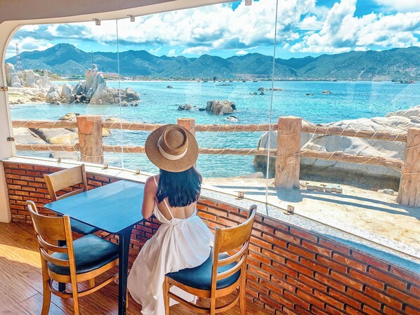 A dining area with a view of a coastal landscape, featuring a table and chairs, a wooden fence, and a stone wall. - Hon Co Ca Na Resort (Ca Na)