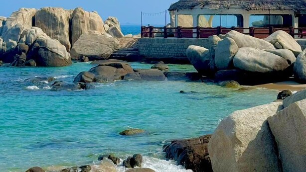 A beach with large rocks, clear turquoise water, and a small building with a thatched roof.